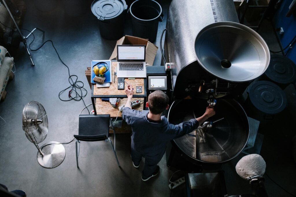 Aerial view of a man working at a coffee roaster with laptop and tools in an industrial workshop setting.